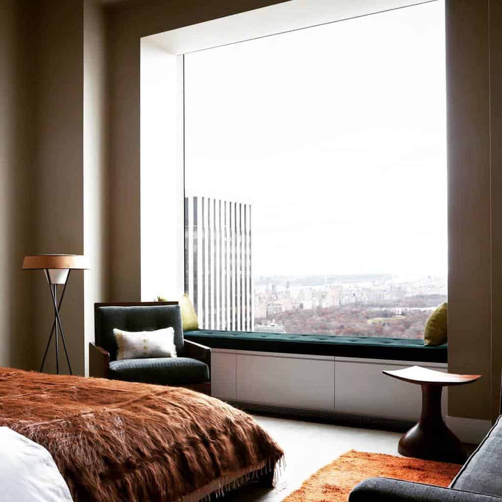 Luxurious bedroom with a floor-to-ceiling bay window, offering a breathtaking cityscape view, cozy seating, and warm, modern decor.