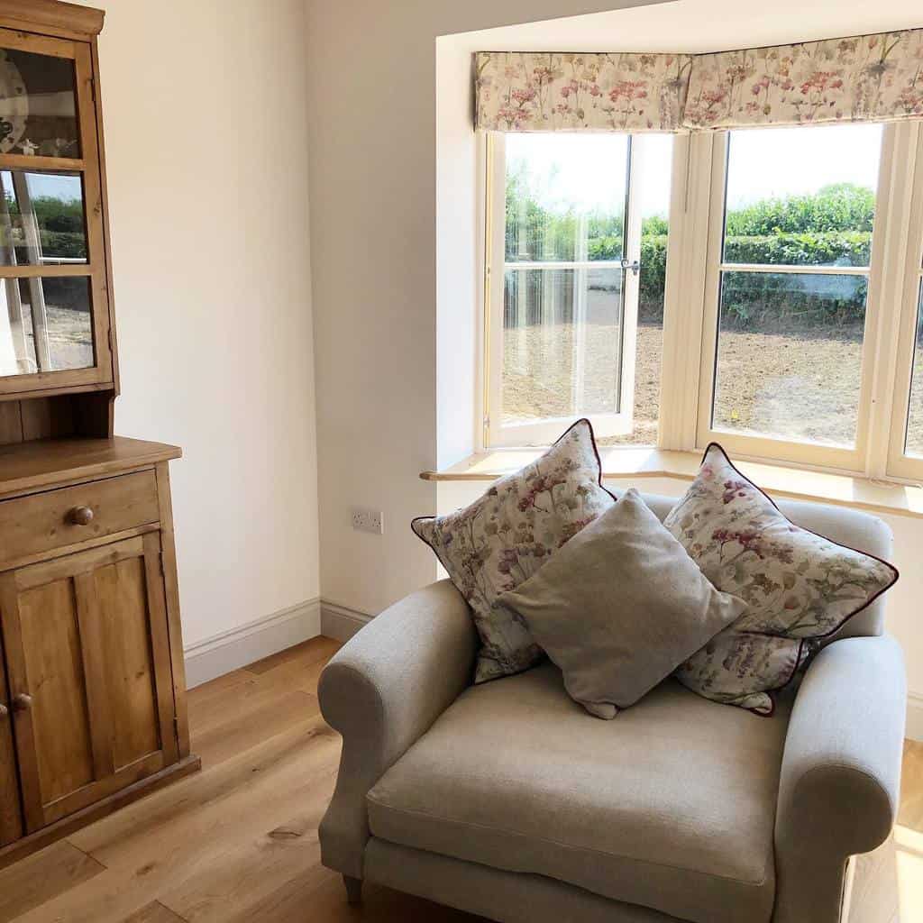 Cozy reading nook with a bay window, floral valance, plush armchair, and soft cushions, creating a charming and relaxing countryside retreat.