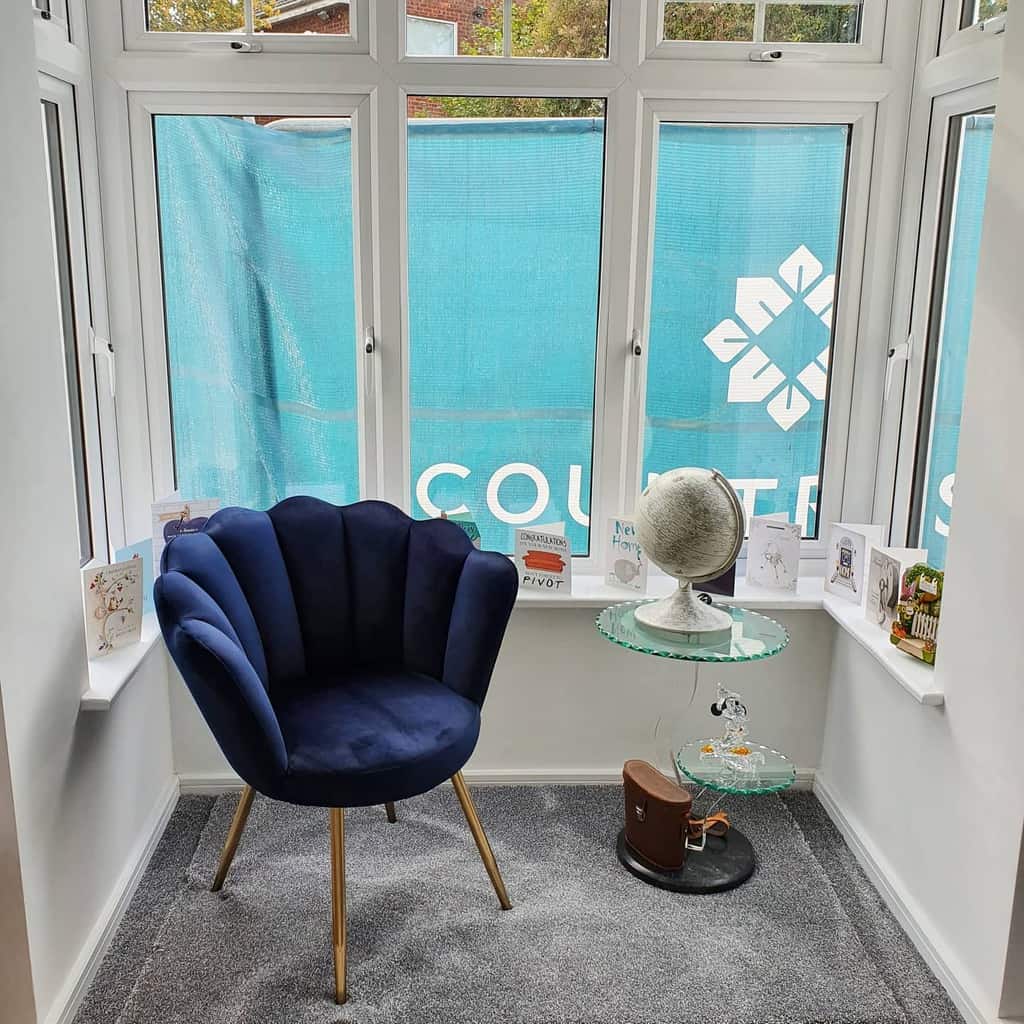 Modern bay window nook with a plush navy blue accent chair, a sleek glass side table, and a cozy carpeted space for relaxation or reading.