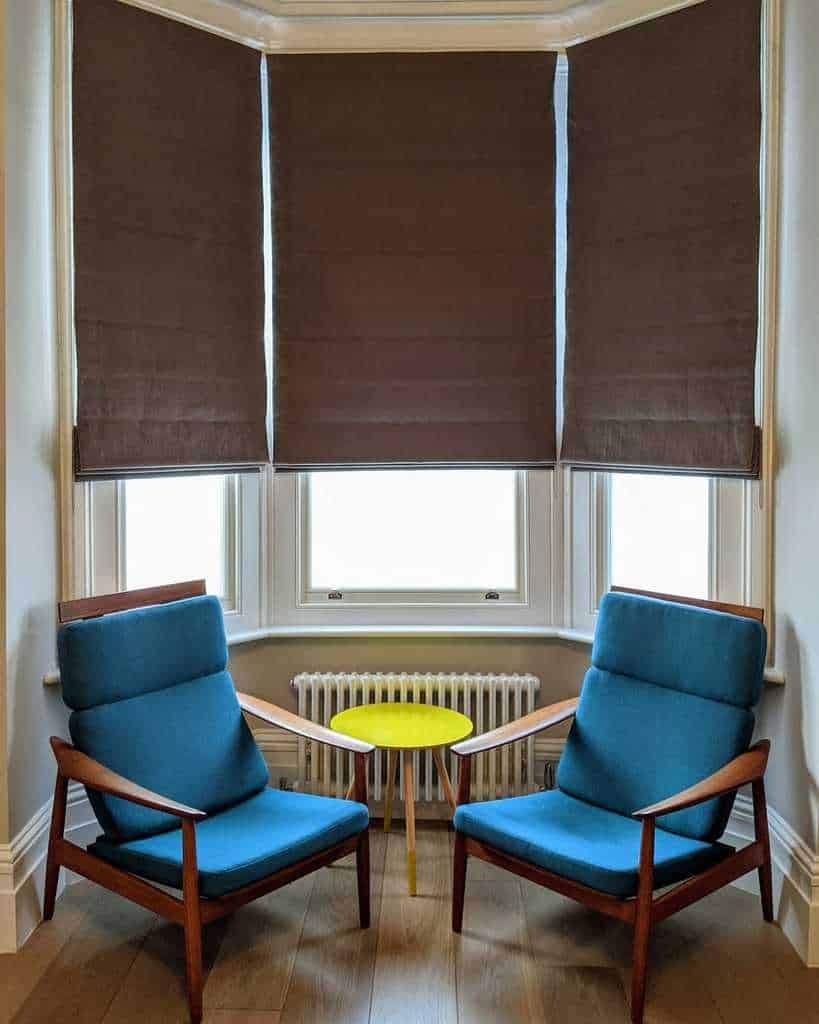 Stylish bay window nook with mid-century modern chairs, bold blue upholstery, dark Roman blinds, and a bright yellow accent table for a cozy touch.