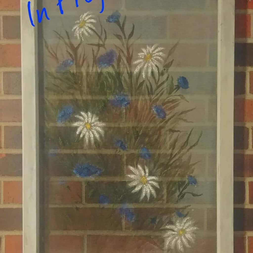 Repurposed old window screen featuring hand-painted wildflowers with white daisies and blue blossoms, set against a brick wall background.