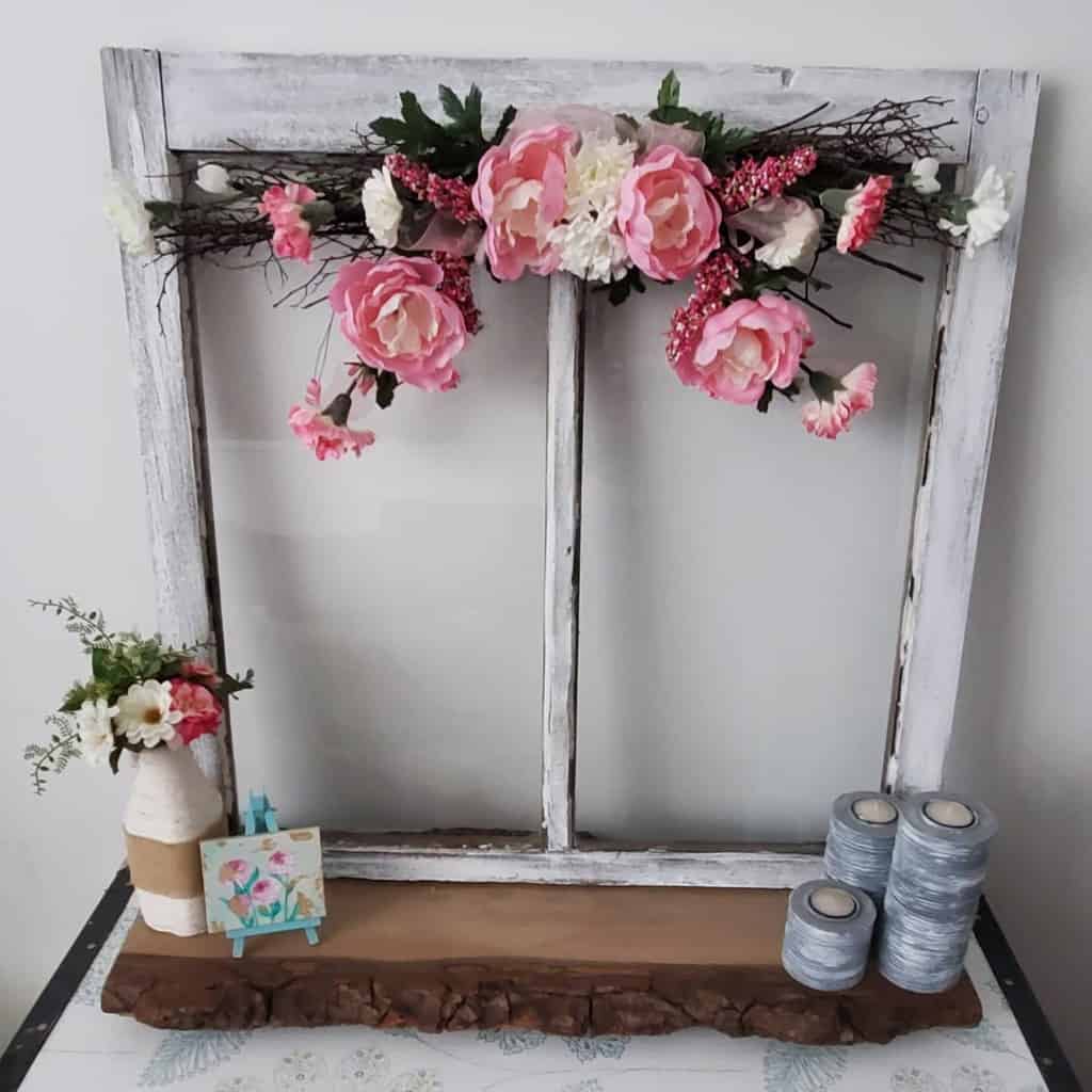 Shabby chic repurposed old window frame with distressed white paint, decorated with pink floral garland and rustic tabletop accents.