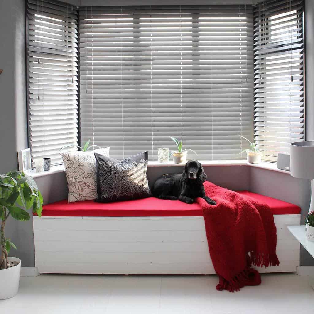 Modern bay window nook with a built-in bench, bold red cushion, cozy throw, and sleek gray blinds, creating a stylish and inviting space.