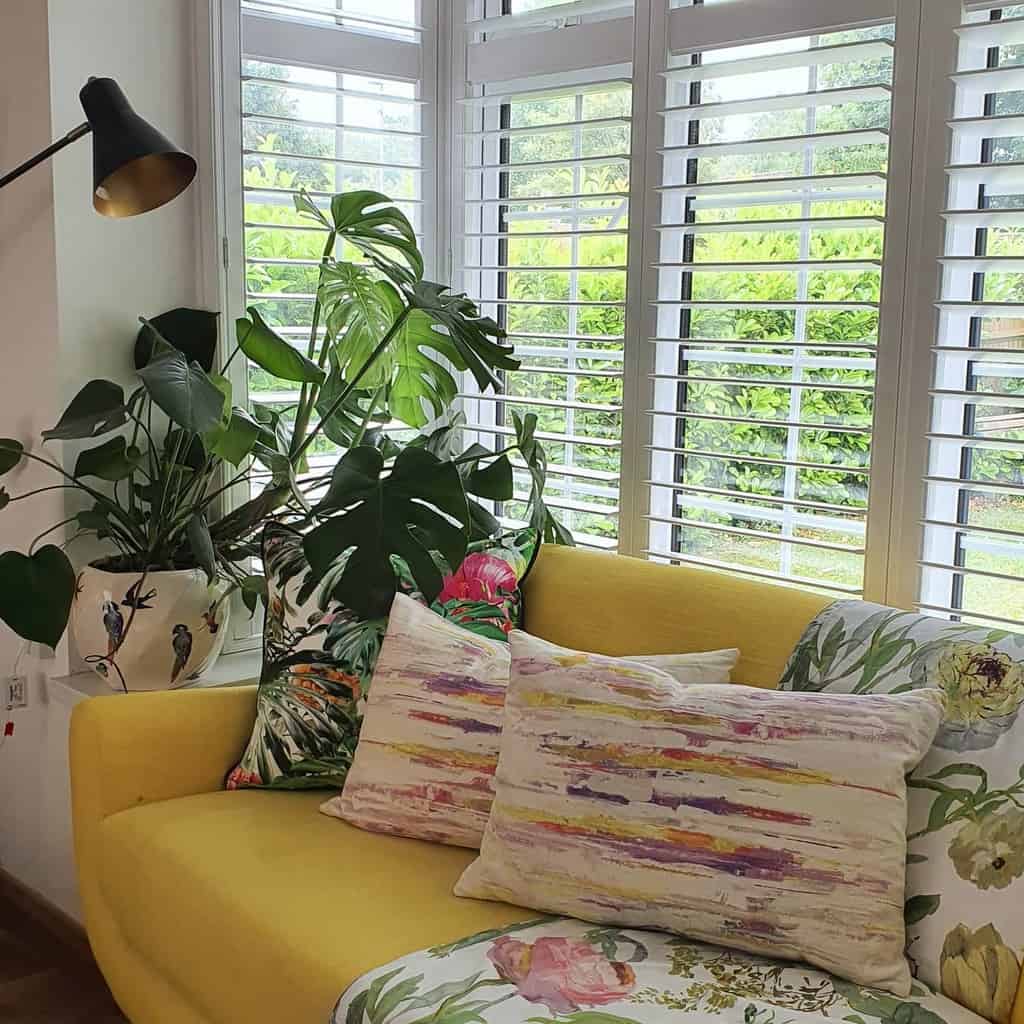 Vibrant bay window nook with white plantation shutters, a bold yellow sofa, lush greenery, and colorful pillows, creating a bright and cozy retreat.