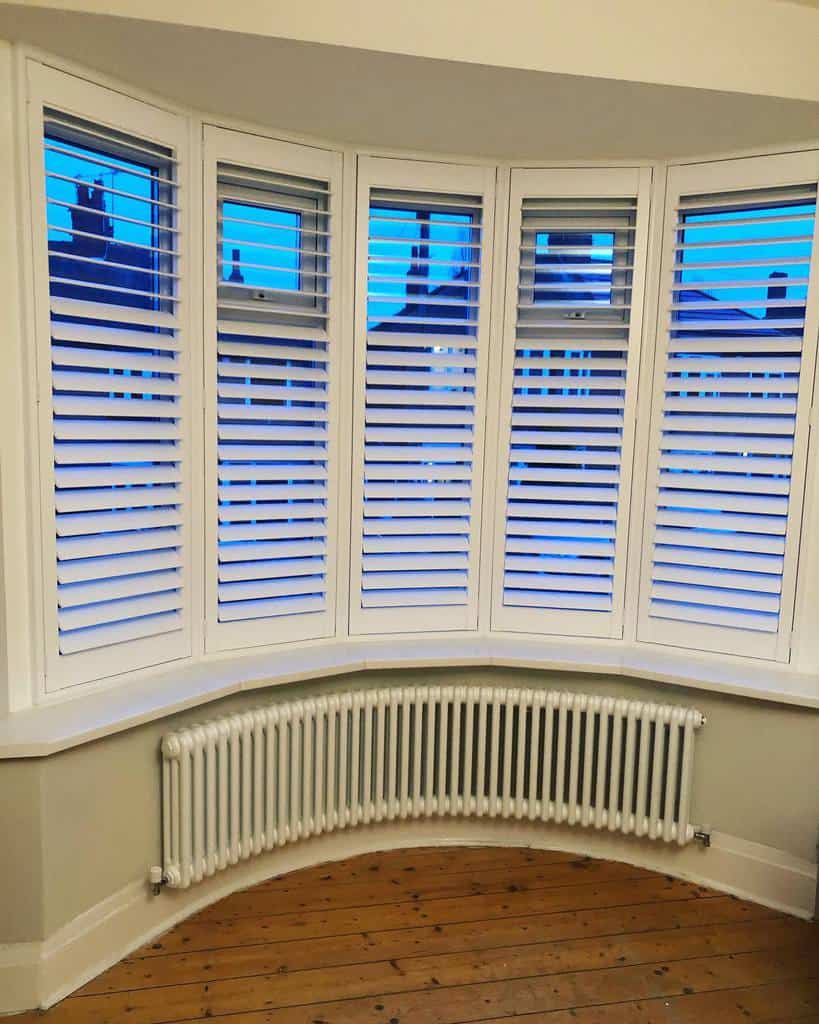 Classic bay window with white plantation shutters, curved radiator, and wooden flooring, offering a bright and elegant look with privacy control.