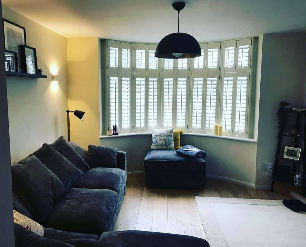 Modern living room with a bay window, white plantation shutters, plush gray seating, and warm lighting, creating a stylish and cozy retreat.