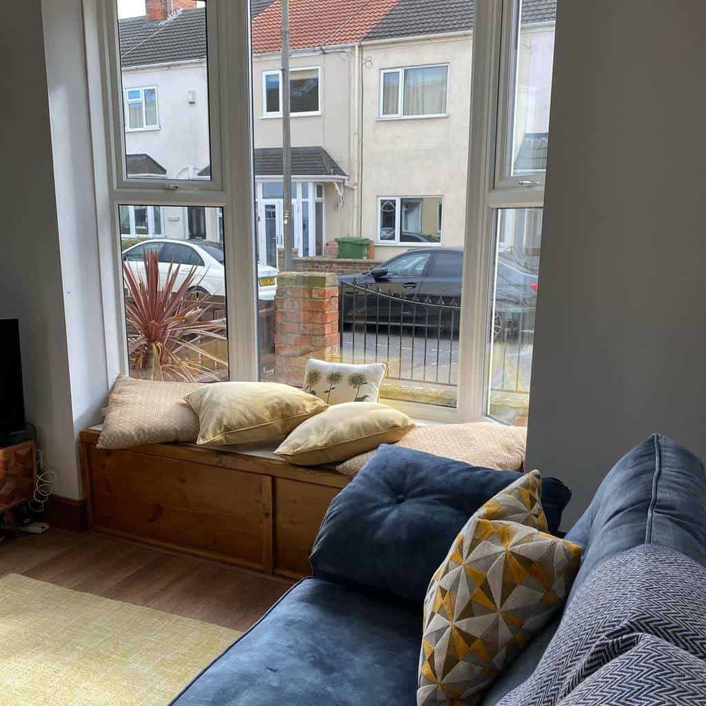 Cozy bay window nook with a wooden storage bench, soft cushions, and a plush blue sofa, creating a warm and inviting living space.