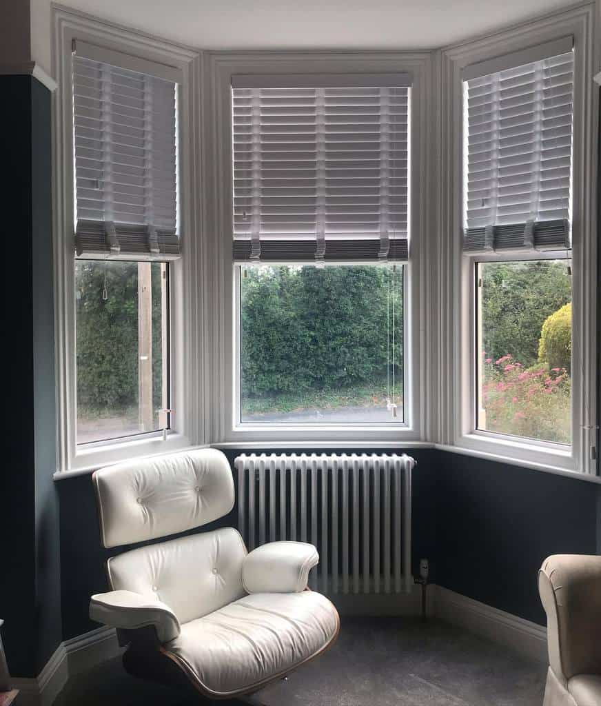 Modern bay window nook with sleek gray blinds, a classic Eames lounge chair, and a cozy radiator, creating a stylish and relaxing reading space.