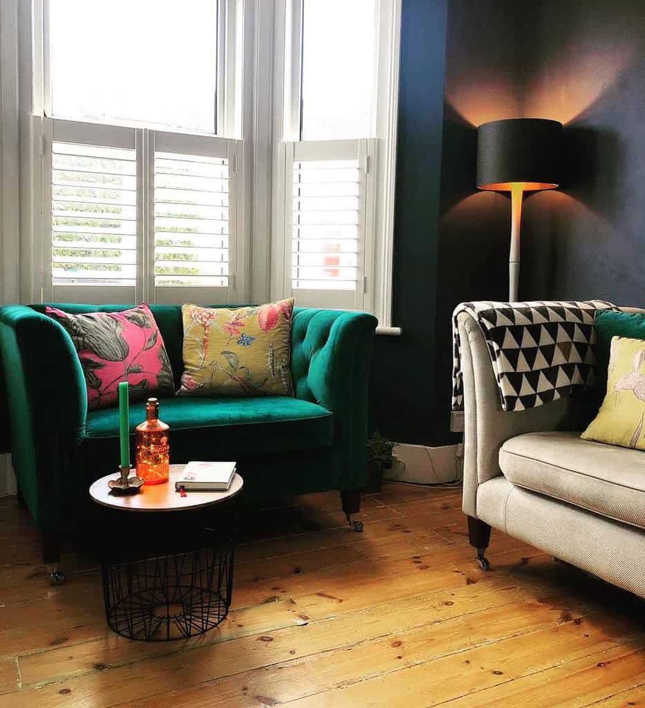 Cozy bay window nook with white shutters, a bold green velvet loveseat, colorful cushions, and warm lighting, creating a stylish and inviting space.