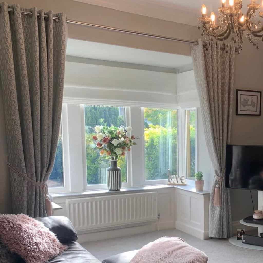 Elegant bay window nook with floor-length drapes, a crystal chandelier, soft neutral tones, and a floral centerpiece, creating a refined and cozy space.