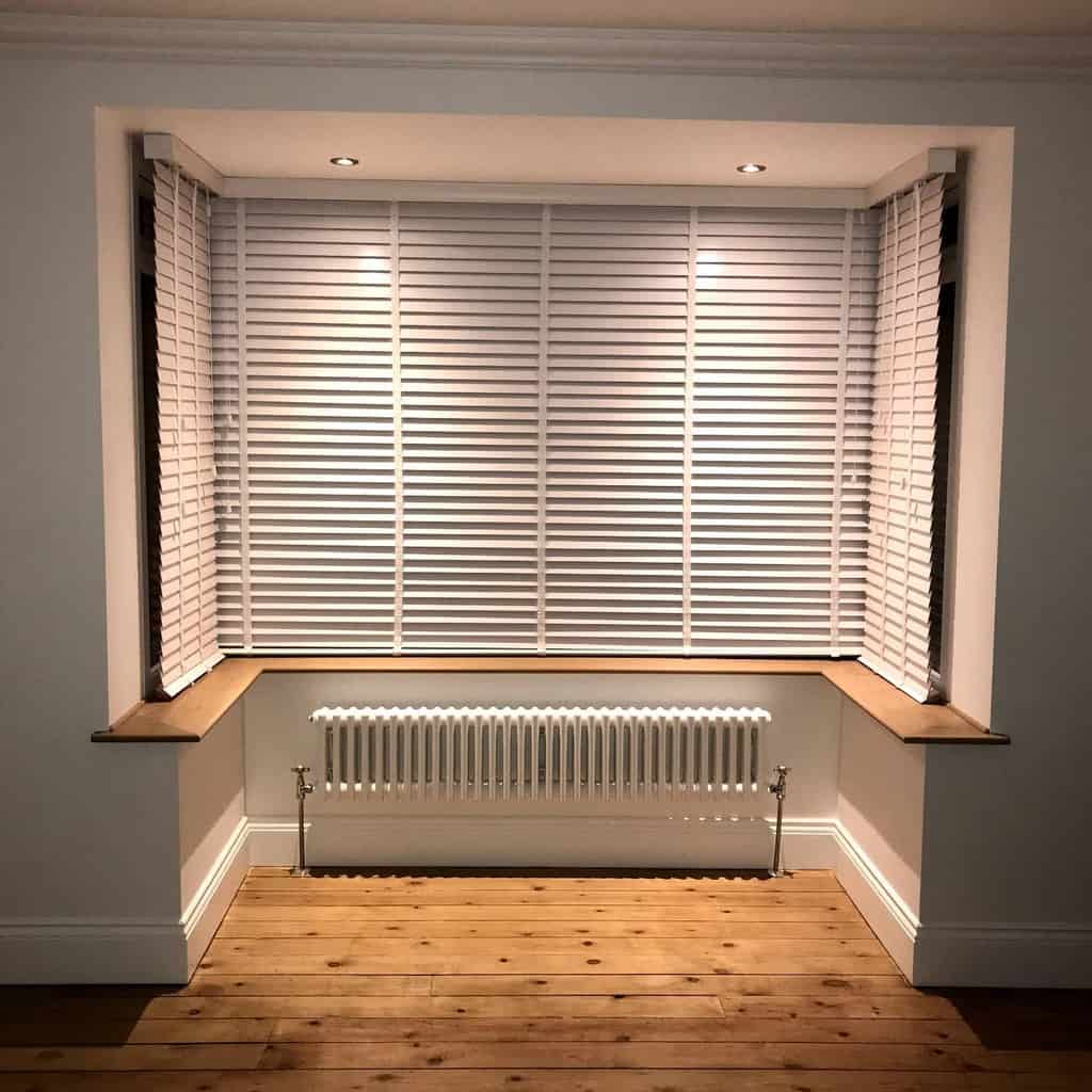 Modern square bay window with sleek white blinds, recessed lighting, wooden ledges, and warm wood flooring, creating a clean and stylish space.