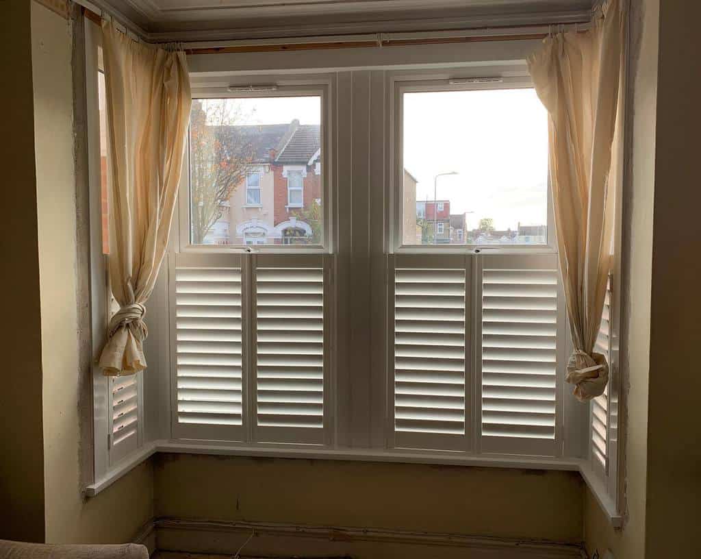 Classic square bay window with white plantation shutters and soft, tied-back curtains, offering privacy while letting in natural light.