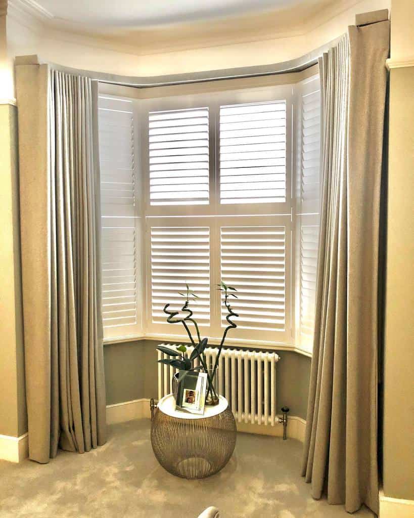 Elegant Victorian bay window with white plantation shutters, floor-length drapes, a stylish radiator, and a modern plant stand for a sophisticated look.