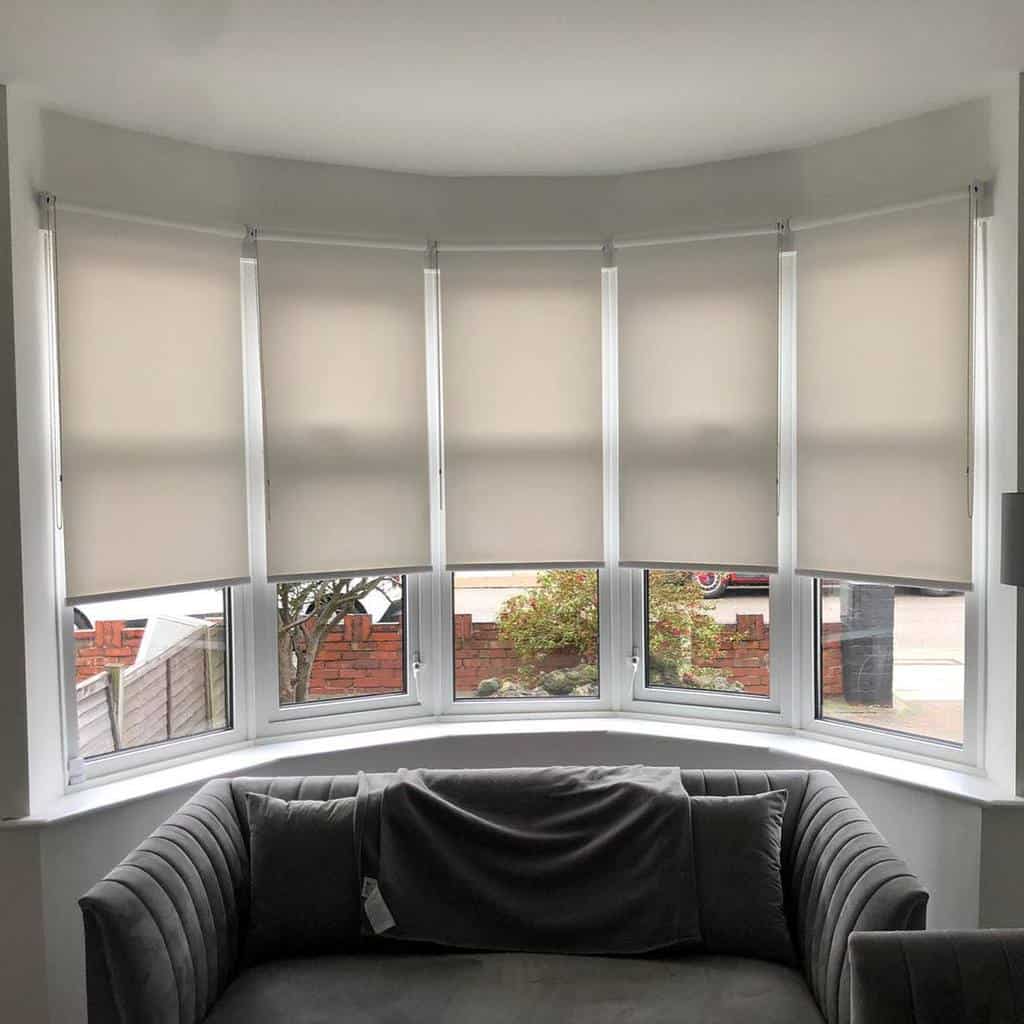 Modern bay window with sleek roller blinds, a curved velvet sofa, and a minimalist design, creating a cozy and stylish living space.