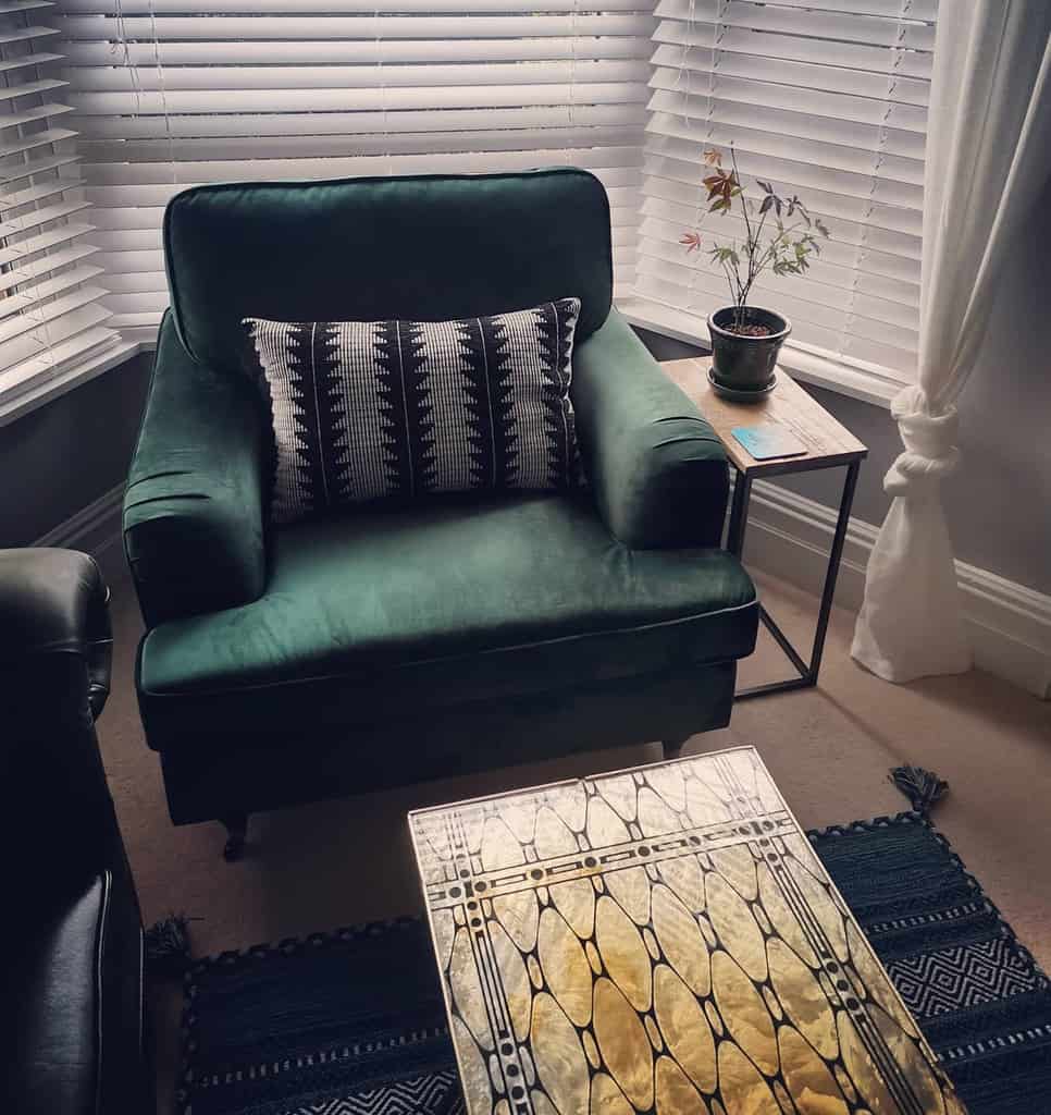 Cozy bay window nook with a plush green velvet armchair, white blinds, a vintage brass table, and warm decor for a stylish and relaxing space.