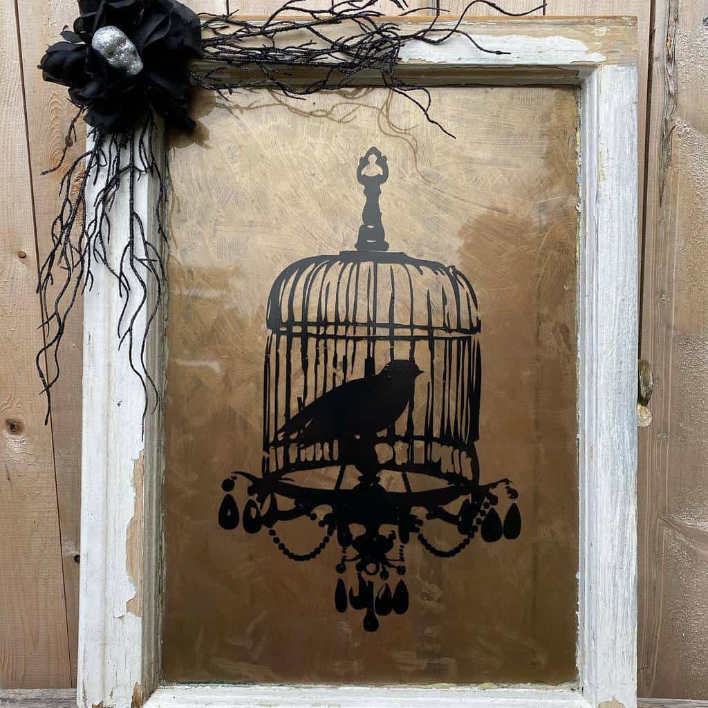 Repurposed old window frame with a gothic-style birdcage silhouette on glass, adorned with black floral and branch accents for a vintage look.