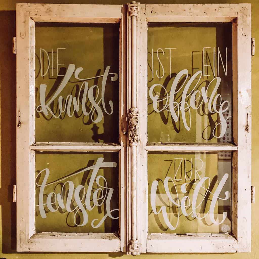 Vintage distressed window frame repurposed as wall art, featuring an elegant hand-lettered German quote on glass about art as a window to the world.