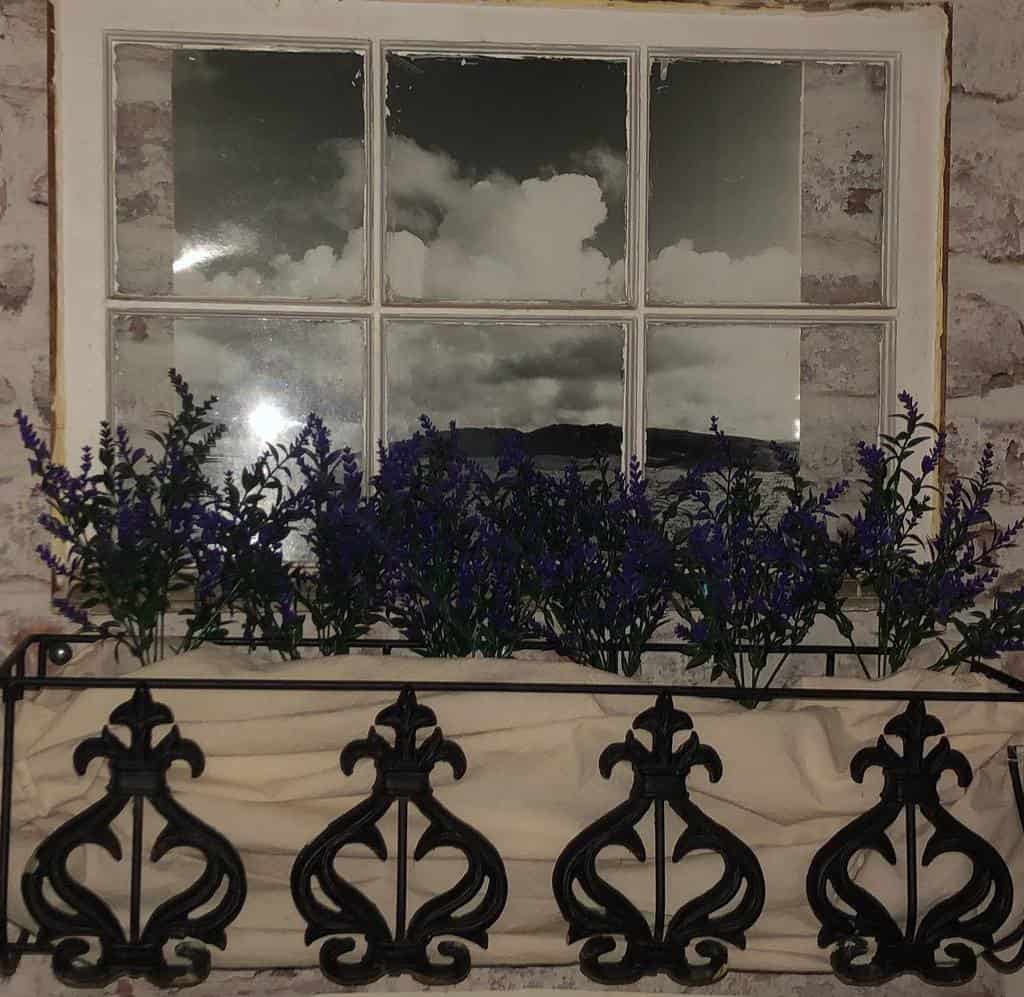 Repurposed old window frame with mirrored glass, styled as a faux balcony scene with an ornate wrought iron planter filled with lavender.