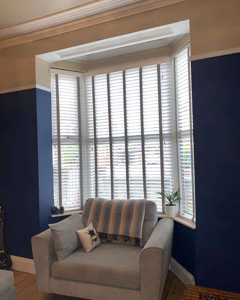 Cozy bay window nook with white blinds, a soft armchair, and deep blue walls, creating a stylish and inviting reading corner.