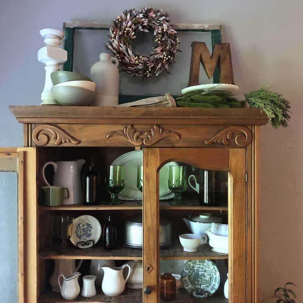 Vintage wooden cabinet styled with a repurposed old window frame, a decorative wreath, rustic pottery, and greenery for a cozy farmhouse touch.