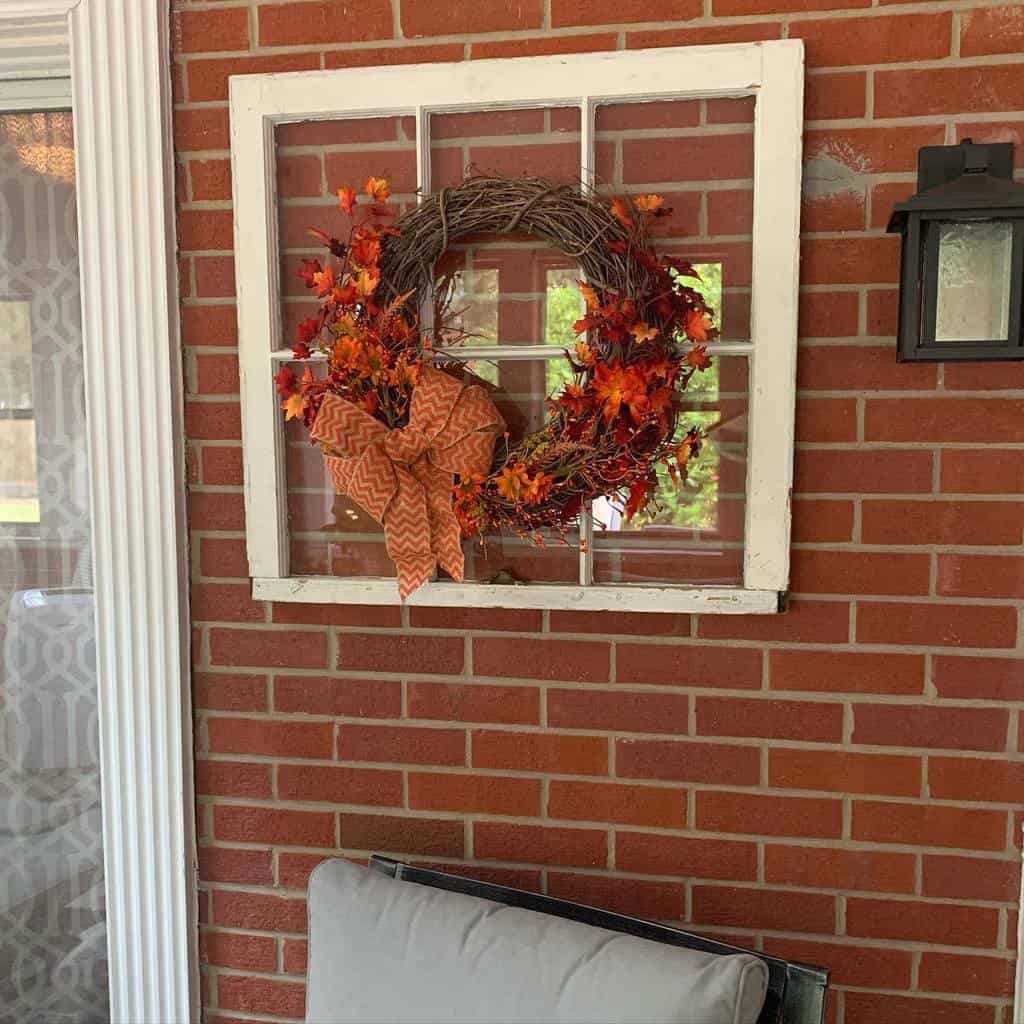 Repurposed old window frame mounted on a brick wall, decorated with a fall-themed wreath featuring autumn leaves, a burlap bow, and warm tones.