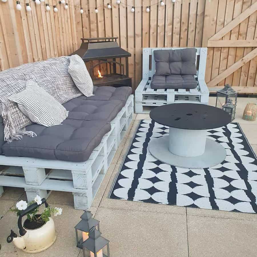 Cozy outdoor pallet seating with gray cushions, a spool coffee table, lanterns, string lights, and a fireplace for a warm ambiance