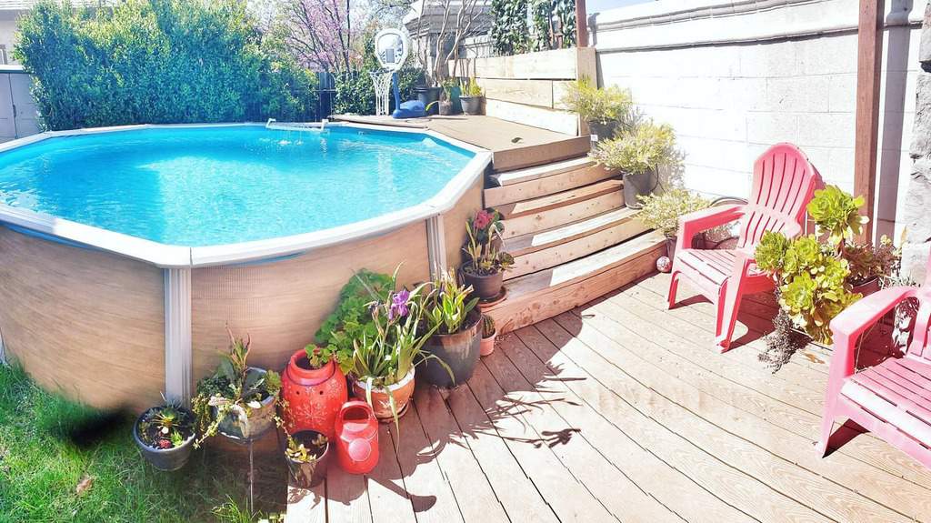 Above-ground pool beside a wooden deck with red chairs and potted plants, steps lead up to a raised seating area