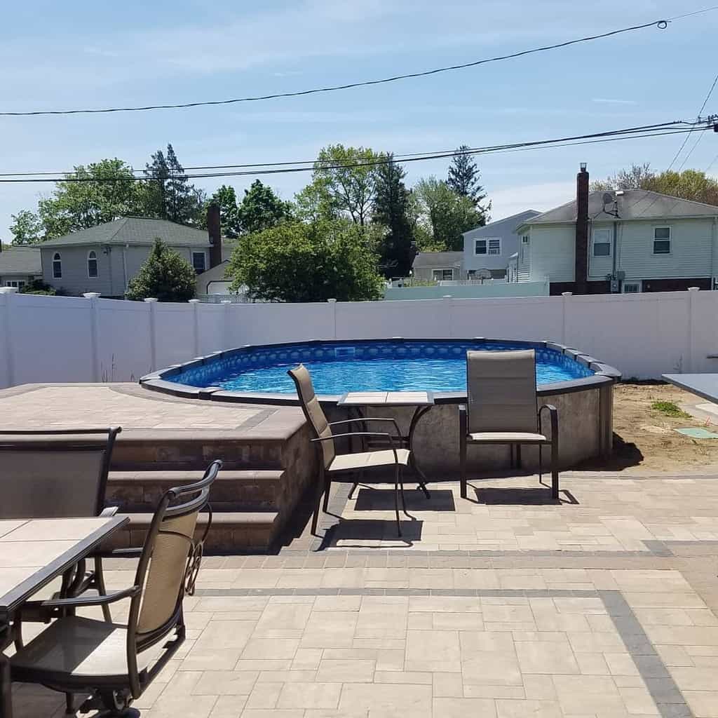 Above-ground pool in a backyard with a stone patio, surrounded by a white fence, with patio furniture including chairs and a table