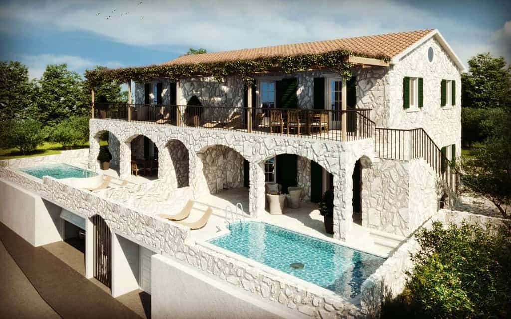 Stone villa with arched balcony, surrounded by trees, features two pools and sun loungers, with a terracotta roof and green shutters