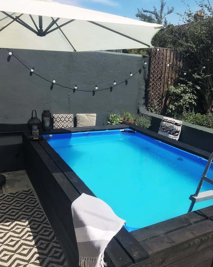 Small backyard pool with black wooden decking, a white umbrella, string lights, and cozy seating on a patterned rug