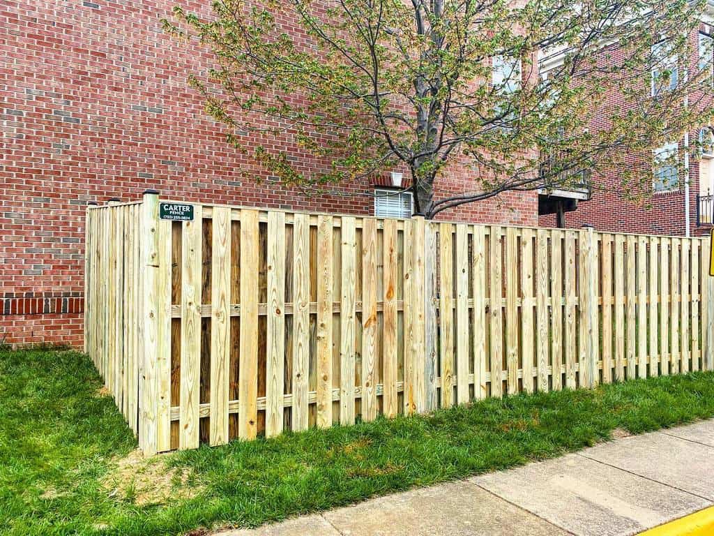 Pallet-style wooden fence enclosing a small yard near a brick building, featuring vertical slats for a rustic and airy look