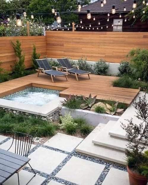 Cozy backyard with a wooden deck, two lounge chairs, a small pool, string lights overhead, and surrounding greenery