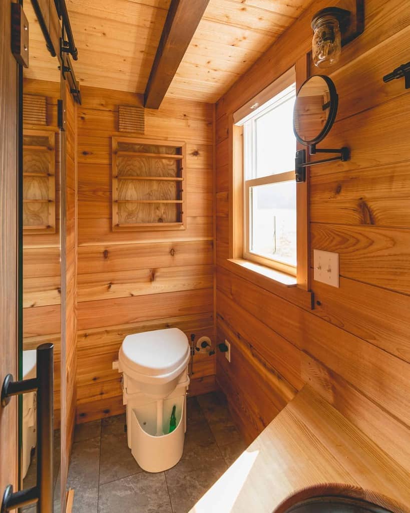 Rustic tiny house bathroom with warm wood paneling, a composting toilet, and a built-in shelf. A large window adds natural light.