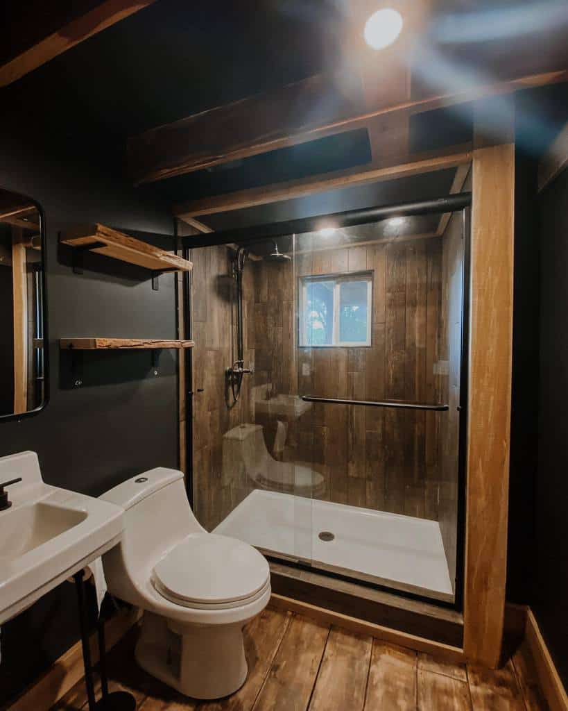 A rustic bathroom with a wooden floor, glass shower, and wall shelves exudes warmth and charm as dim lighting enhances the cozy atmosphere in this small space design.