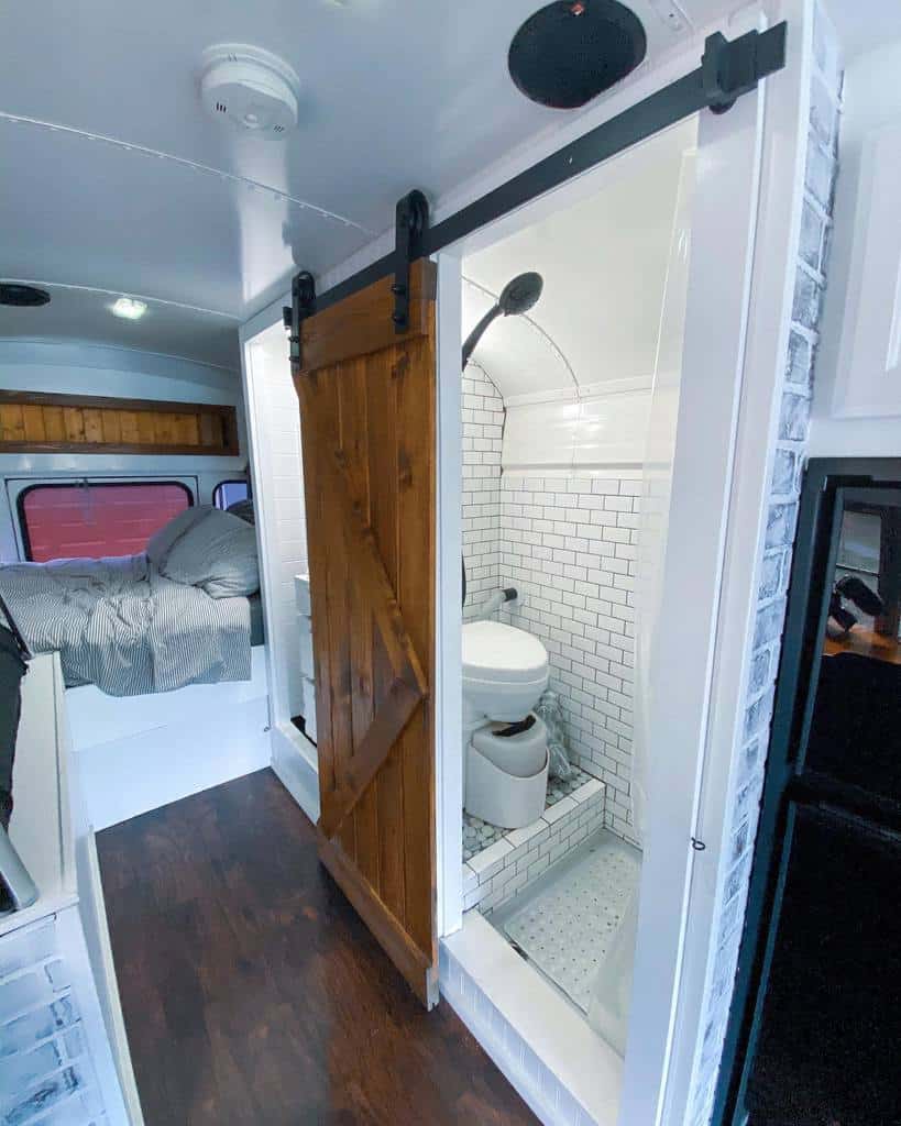 A converted bus interior showcases small spaces design ideas with a rustic wooden sliding door separating a subway-tiled bathroom from the cozy sleeping area.