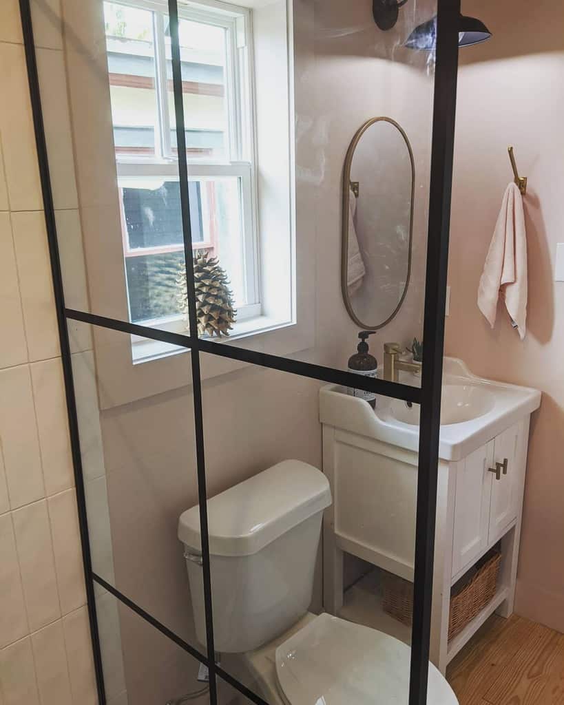 Small bathroom with a modern black-framed glass shower, white vanity, and soft pink walls. A brass mirror and fixtures add warmth.