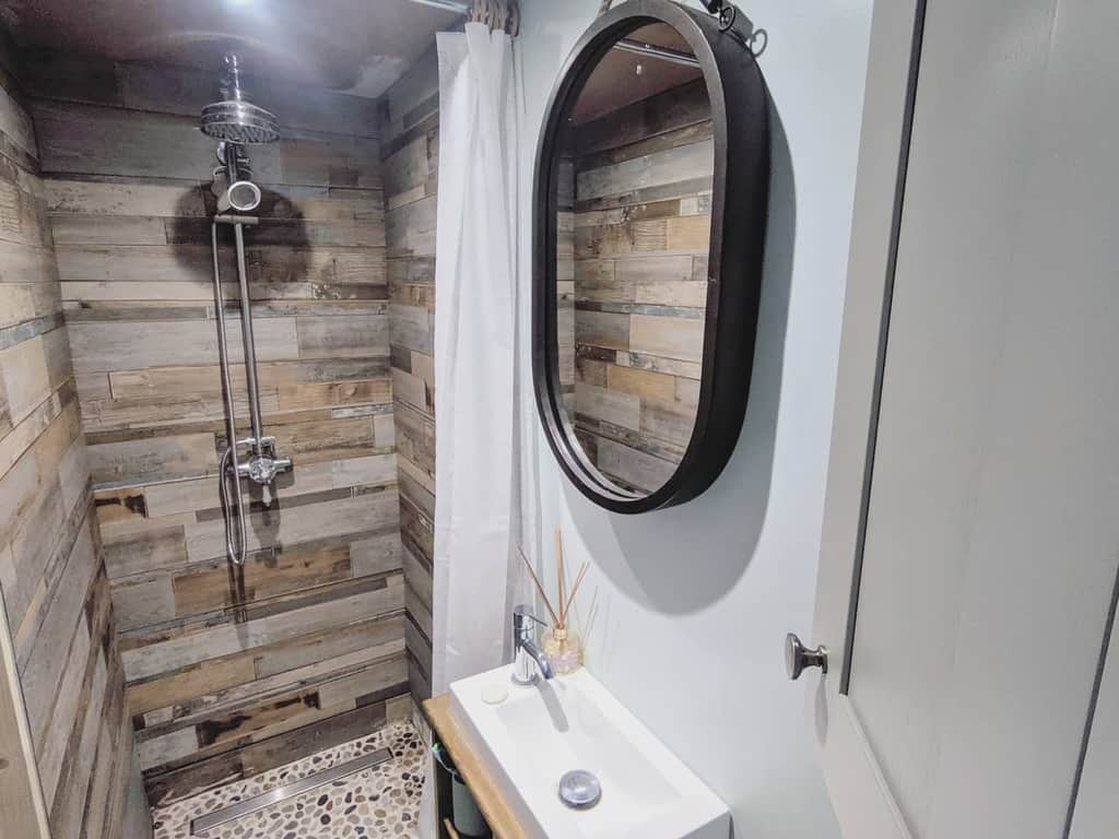 Rustic tiny house bathroom with wood-paneled shower, pebble flooring, and a compact sink. A black oval mirror adds contrast.