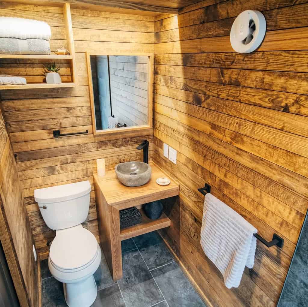 A rustic bathroom showcases small spaces design ideas with wood-paneled walls, a stone sink on a wooden vanity, open shelves, and a towel rack.