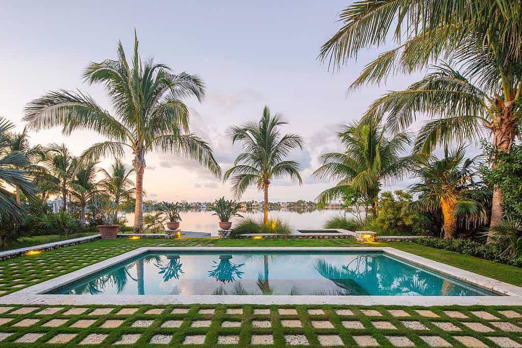 A tropical backyard with a square pool, surrounded by palm trees and grass, overlooking a serene body of water at sunset