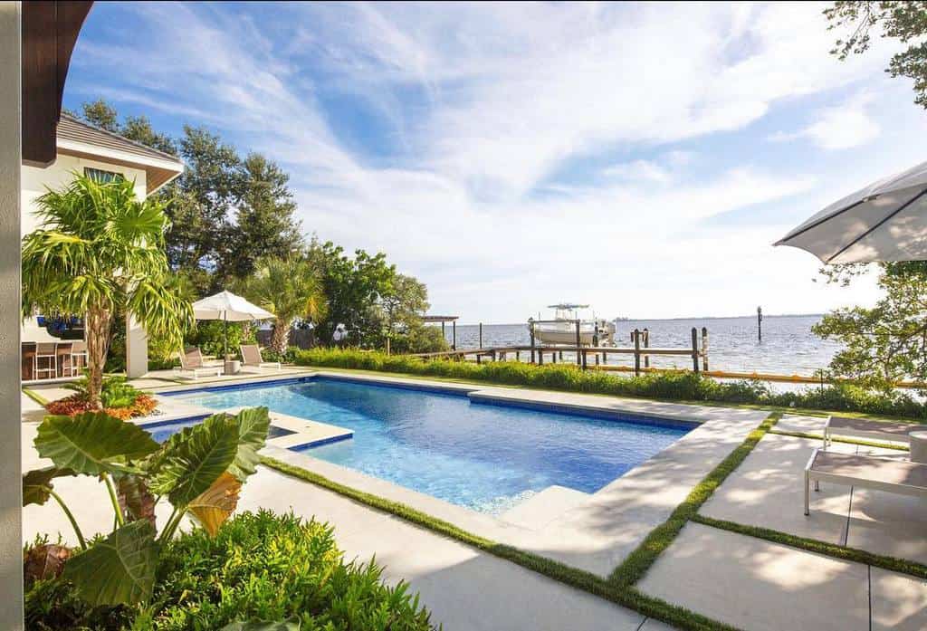 Sunny backyard with a clear pool, tropical plants, and a view of the waterfront with a dock and boat in the background