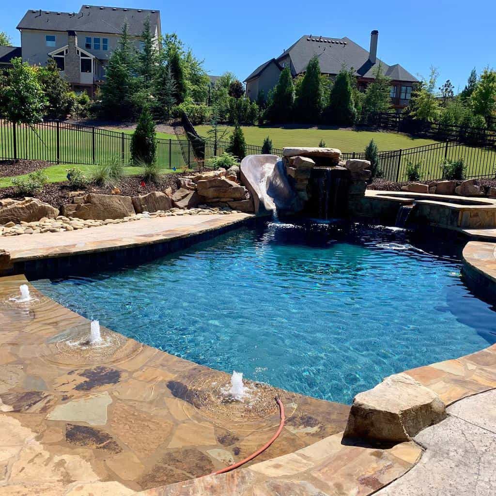 Backyard pool with a rock waterfall feature and slide, surrounded by stone patio and landscaped greenery, near houses and trees