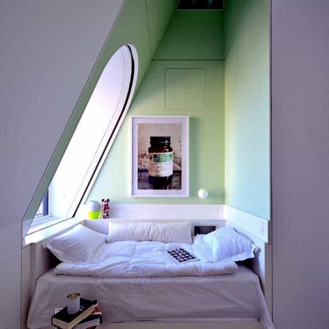 Cozy alcove bedroom with a green accent wall, white bedding, angular window, and artwork above the bed.