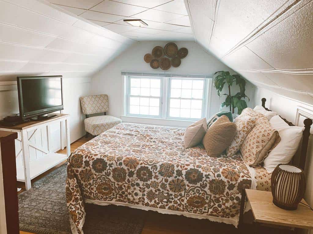 Cozy attic bedroom with a vintage quilt, wooden accents, and soft lighting. A large window brightens the space, creating an inviting retreat.