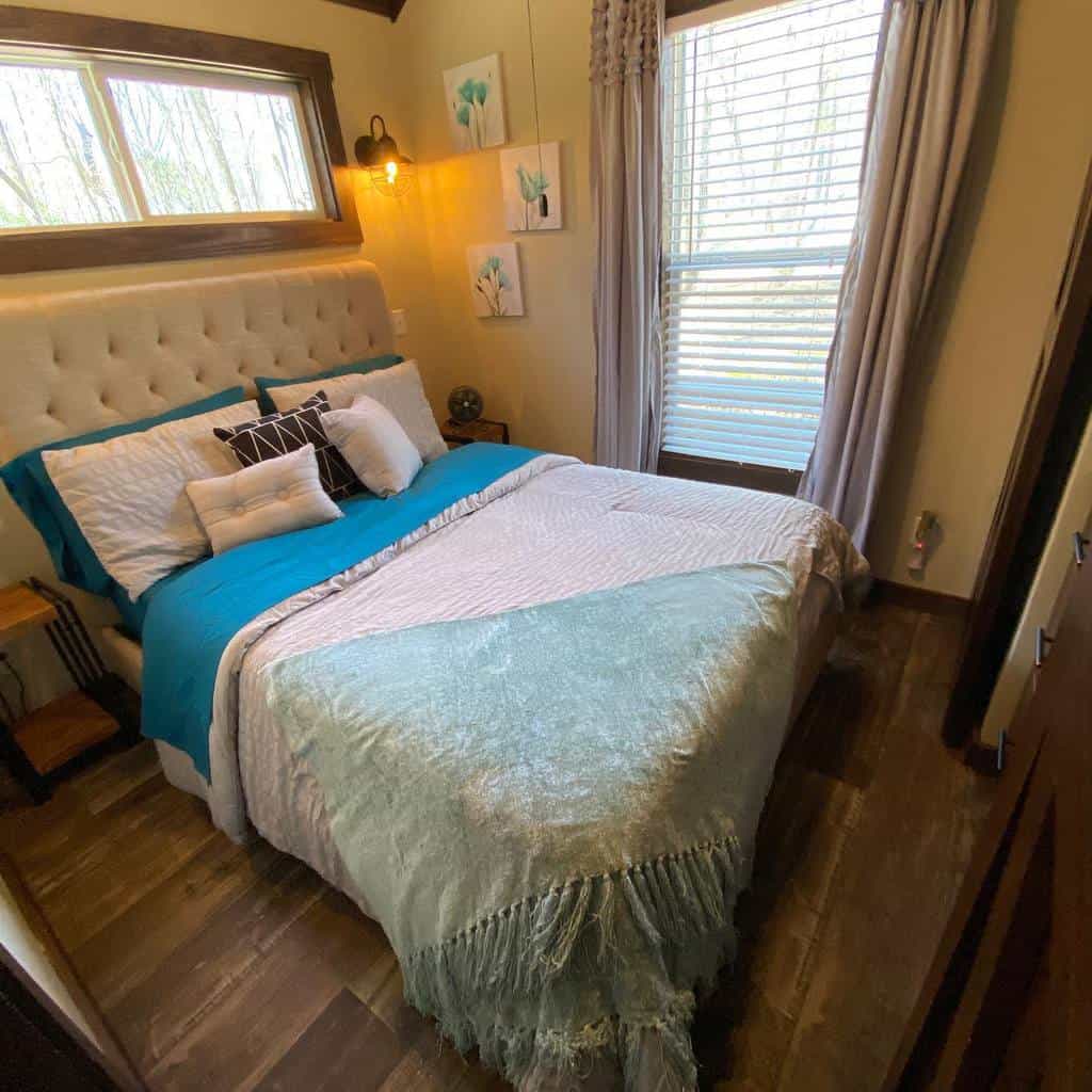 Cozy tiny house bedroom with a tufted headboard, soft blue bedding, and warm lighting. Large windows bring in natural light and a serene view.