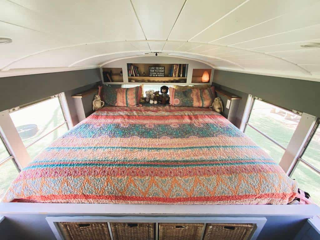 Cozy bus-converted bedroom with a colorful quilt, built-in bookshelves, and storage baskets. Large windows bring in natural light.