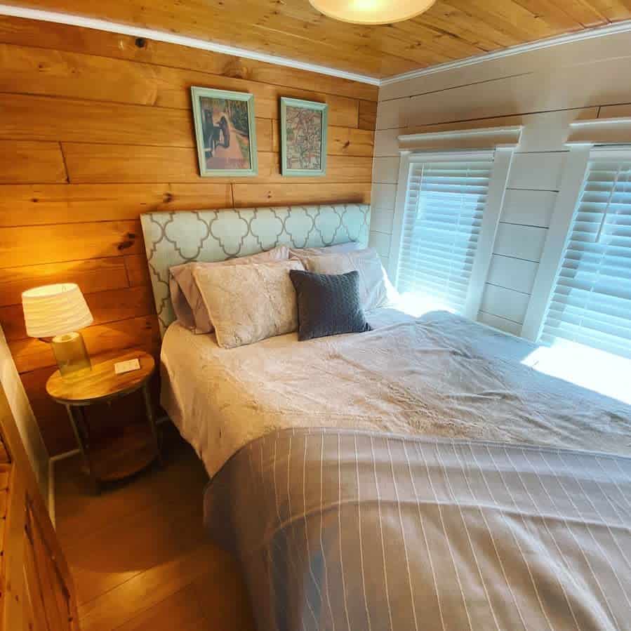 Small bedroom with a wooden accent wall, bed with patterned headboard, side table with lamp, and two framed pictures.