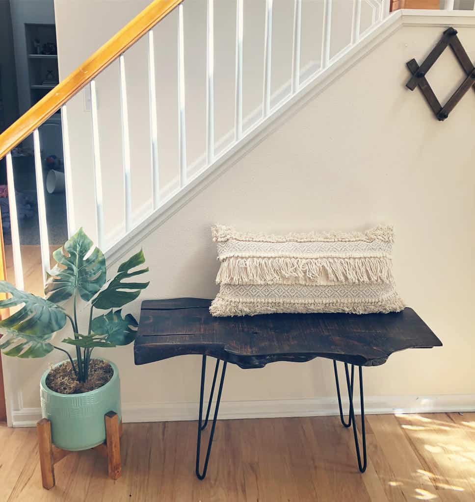 Boho-style entryway with a dark wood live-edge bench, textured fringe pillow, potted plant, and a cozy nook under the staircase