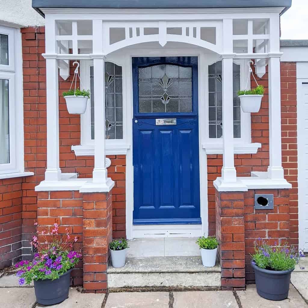 A house entrance with a vibrant blue door, white porch, hanging and potted plants, set against red brick walls