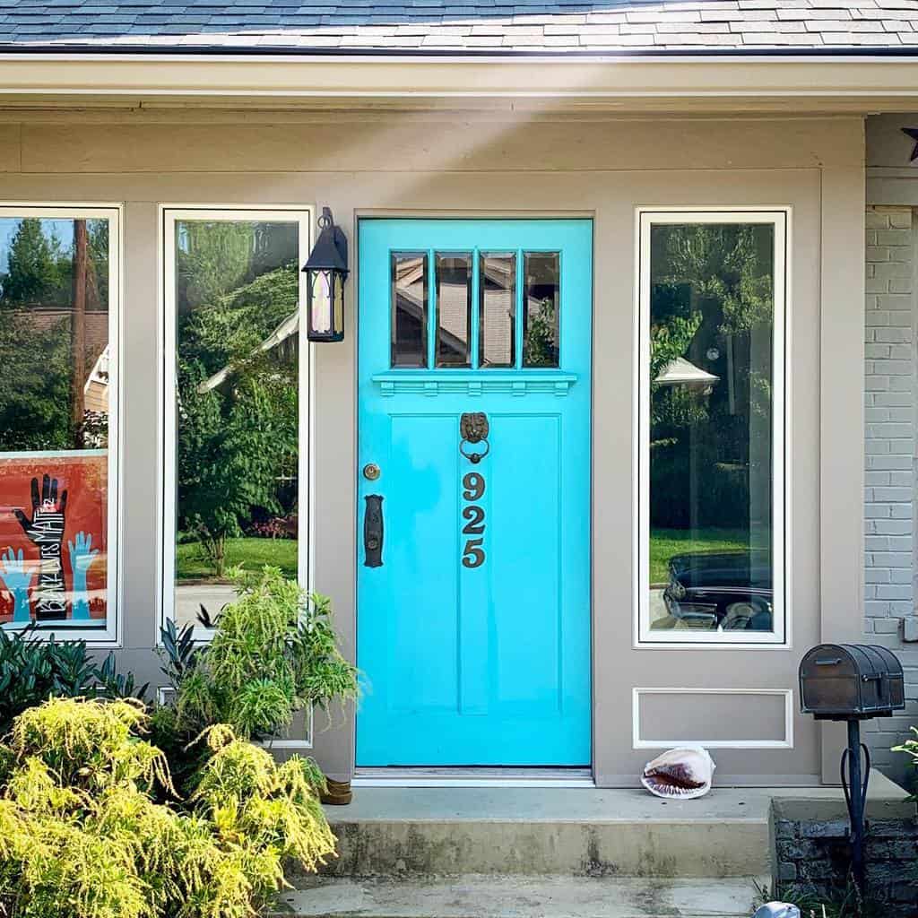 Bright turquoise door with brass knocker and numbers 925, flanked by windows, surrounded by plants and a small mailbox on the side