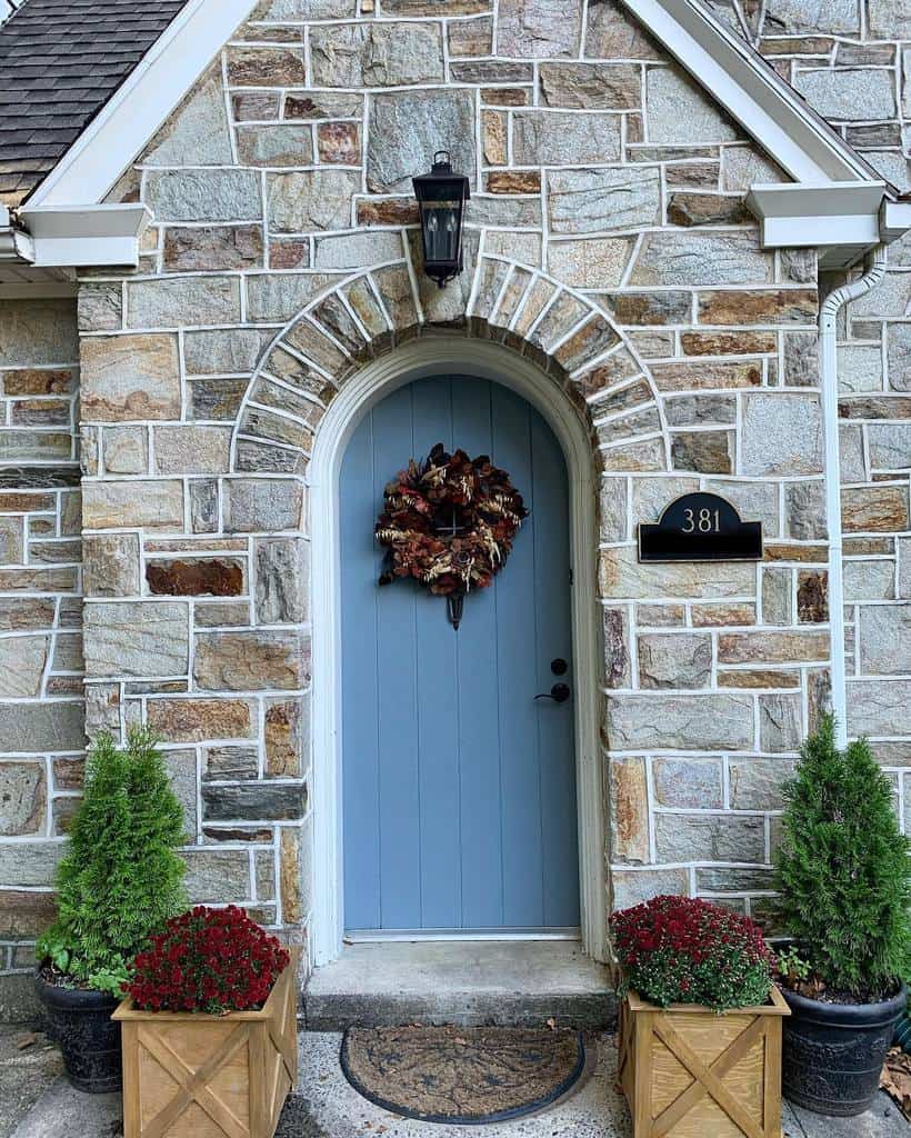 Charming stone house entrance with a light blue door, autumn wreath, lantern, and number plaque, flanked by potted plants and flowers